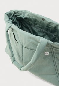 Τσάντα tote από ανοιχτό πράσινο ύφασμα με επενδυμένες λαβές, ανοικτό πάνω μέρος που δείχνει εσωτερική τσέπη με φερμουάρ και εξωτερική μπροστινή τσέπη με μικρή ετικέτα.