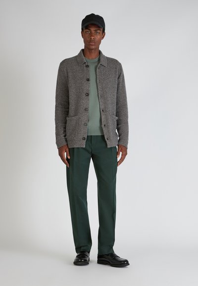 Grauer Wollcardigan mit Knopfverschluss und zwei Taschen, kombiniert mit einem hellgrünen Pullover und dunkelgrünen Hosen. Schwarze Mütze und Schuhe.