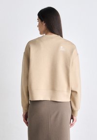 Sudadera corta beige con mangas largas, puños y dobladillo acanalados, que presenta un pequeño logo en la parte superior de la espalda. La modelo lleva una falda marrón.