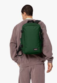 Grön tygryggsäck med rektangulär form, sidofickor och justerbara axelremmar. Har dragkedjor och en logopatch på framsidan.