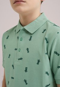 Grön polotröja med små mönster av fyrar, korta ärmar, krage och tre gröna knappar. Texturerad tyg med andningsbar finish.
