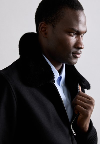 Svart ullblandad jacka med krage av konstfuskpäls. Har en silverfärgad dragkedja och ribbade manschetter. Bärs över en ljusblå skjorta.