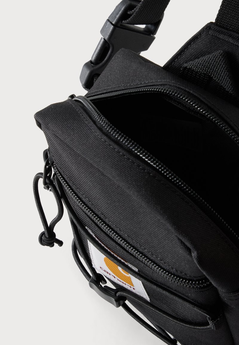 Black shoulder bag with open zippered main compartment, adjustable strap with buckle, and front logo patch and cord detail.