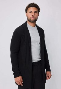 Homme aux cheveux courts et bouclés portant une chemise gris clair, un cardigan noir ouvert et un pantalon noir, debout devant un fond clair uni.