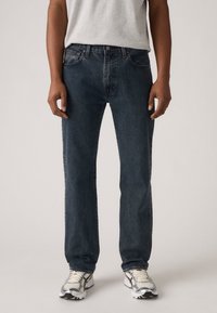 Donkerblauwe denim jeans met een rechte pasvorm, vijf zakken en subtiele slijtage details. G gedragen met een grijze t-shirt en sportschoenen.