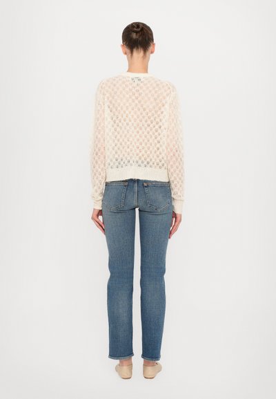 Cardigan di pizzo color crema con maniche lunghe e orlo a coste, abbinato a jeans blu aderenti e ballerine beige, visto da dietro.