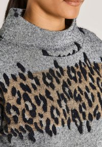 Nahaufnahme einer Person, die einen grauen Rollkragenpullover mit einem braun-schwarzen Leopardenmuster auf der Brust trägt.