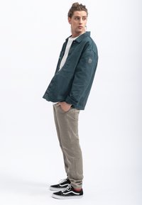 Camicia overshirt verde scuro in cotone, con colletto a punta, tasche e un'etichetta con logo. Abbinata a pantaloni beige chiaro a gamba aderente e sneakers nere.