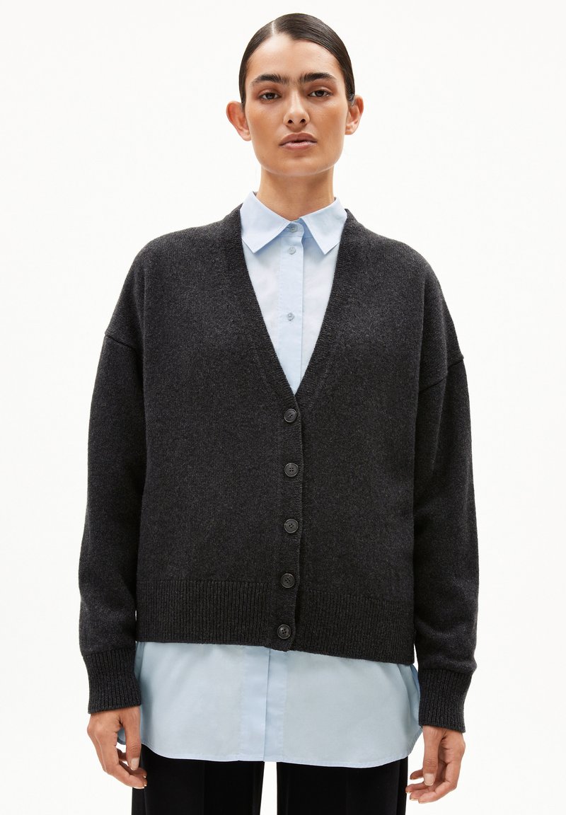 Donkergrijze cardigan met een V-hals, lange mouwen en zes knoopsluitingen. Geribde manchetten en zoom; gedragen over een lichtblauw overhemd met kraag.