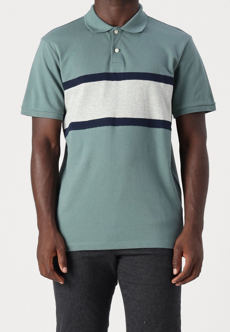 Homme portant un polo vert avec une bande horizontale gris clair bordée de bandes bleu foncé sur la poitrine.
