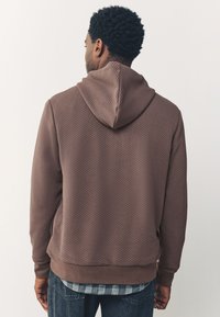 Felpa marrone con cappuccio a coulisse, fondo orlato e maniche lisce, indossata sopra una camicia a quadri.