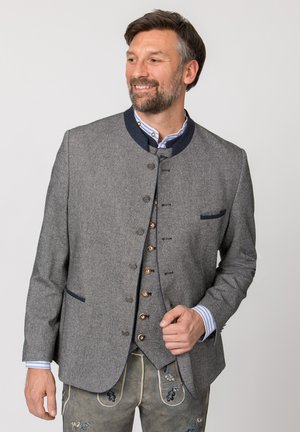 Mann trägt traditionelle graue bayerische Jacke und bestickte Lederhose, lächelt und schaut seitlich vor einfarbigem Hintergrund.