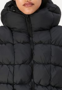 Melna pūkainā jakas ar augstu apkakli, quilted dizainu, priekšējo rāvējslēdzēju un gludu virsmas tekstūru.