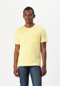 Gul bomulds T-shirt med rund hals og korte ærmer, med en brystlomme og en lille logo detalje. Båret med blå jeans.