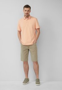 Jasno brzoskwiniowa koszulka polo z kołnierzykiem, zestawiona z beżowymi szortami. Model w zielonkawych trampkach, stojący na jednolitym szarym tle.