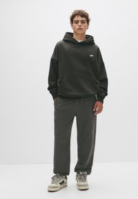 Donkergrijze hoodie met een voorzak en logo op de borst, gecombineerd met bijpassende broek. Model draagt lichtgewicht sneakers met accenten.