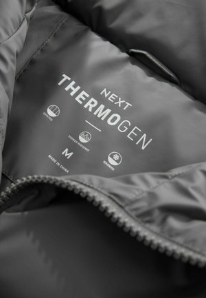 Graue gesteppte Jackeninnenseite mit einem Etikett, auf dem "Next Thermogen" steht. Enthält Symbole für gesteppte Verarbeitung, wasserabweisend, Wärme, Größe M und hergestellt in China.