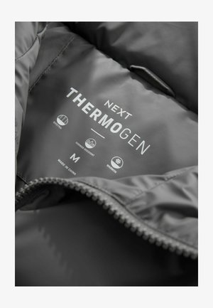 Grå quiltet jakke indvendig med et mærke, der læser "Next Thermogen." Viser ikoner for quiltet, vandafvisende, varme, størrelse M og fremstillet i Kina.