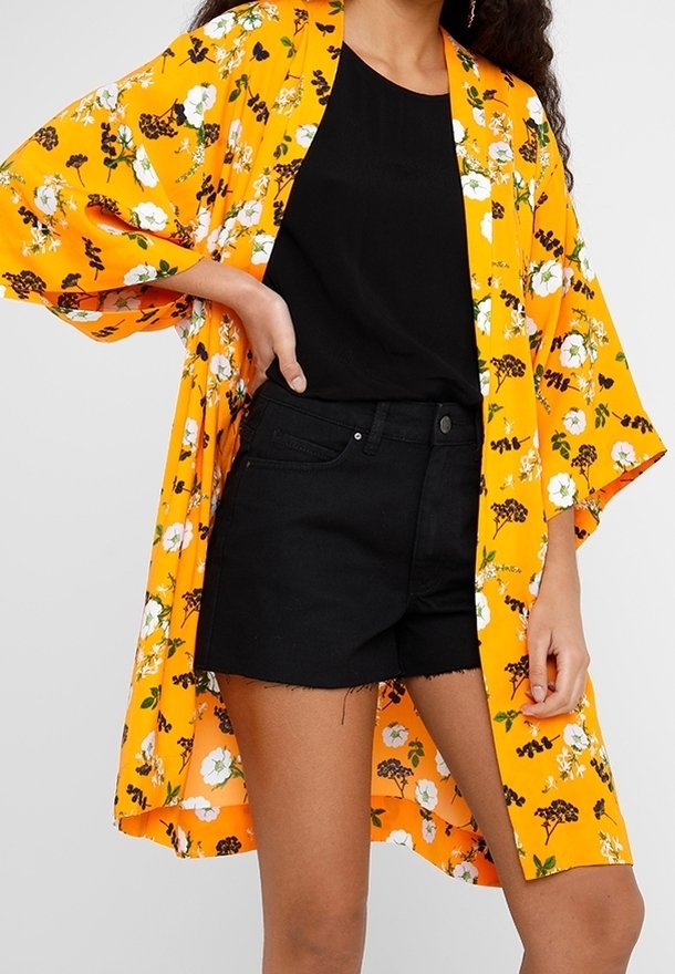Femme portant un kimono à fleurs jaune vif par-dessus un t-shirt noir et un short en jean noir, posant avec une main sur la hanche.