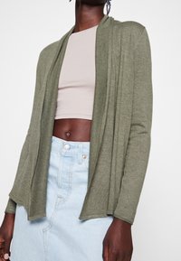 Oliivinvihreä kardigaani, joka on valmistettu kevyestä kankaasta, avoimen edessä olevan muotoilun ja pitkien hihojen kanssa, kerroksittain vaaleanpunaisen lyhyen paidan ja farkkuhameen päällä.