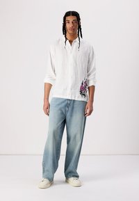 White linen shirt with floral embroidery in pink and black, paired with light blue wide-leg jeans and cream sneakers.