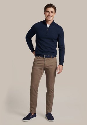 Lachende man staat en draagt een marineblauwe trui met kwartritssluiting, wit ondershirt, bruine broek, marineblauwe riem en marineblauwe instapschoenen op een beige achtergrond.