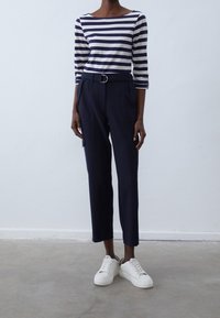 Marinblå smalpassade byxor med bälte i midjan, som matchas med en vit och marinrandig långärmad topp. Vita sneakers fulländar outfiten.