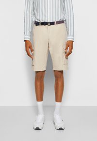 Pantaloni cargo beige con tasche laterali, indossati con una camicia a maniche lunghe a righe blu chiaro e bianco, e sneakers bianche con dettagli neri.