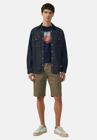 Dunkelblaue Jeanshemd über einem navyblauen Grafik-T-Shirt, khakifarbene Shorts und helle Sneaker mit strukturierten Sohlen. Zwei Brusttaschen am Hemd.