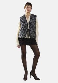 Czarna pikowana kamizelka z białym wzorem florystycznym, założona na kremowy sweter i czarną mini spódniczkę z tekstury, połączona z przejrzystymi czarnymi rajstopami i obcasami.