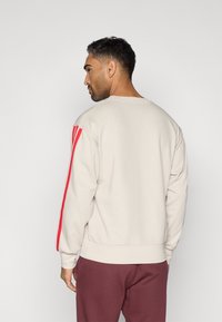 Άντρας που κοιτάζει πλευρικά, φορώντας ένα μπεζ φούτερ με κόκκινες ρίγες στα μανίκια και μπορντό παντελόνι, σε απλό ανοιχτό φόντο.