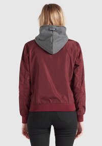 Bordová bomber bunda se šedou kapucí, s elastickými manžetami a pasem, hladkou strukturou a štítkem s logem na zadní straně kapuce.