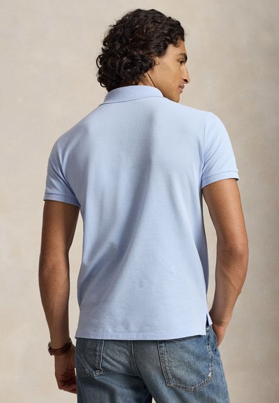 Homme aux cheveux bouclés portant un polo bleu clair à manches courtes et un jean bleu, debout, dos au spectateur.
