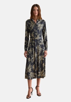 Mujer con vestido midi negro con patrones florales dorados tipo paisley, mangas largas, frente con botones y sandalias negras de tacón alto.