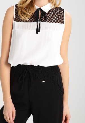 Biała bluzka bez rękawów z czarnym siateczkowym karczkiem, mała kokarda przy kołnierzyku, w zestawie z czarnymi spodniami z koronkowymi detalami i ściągaczem w talii.