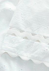 Tissu blanc avec des motifs brodés, des bordures festonnées et des détails en broderie anglaise. Texture douce avec une subtile profondeur et des designs complexes.