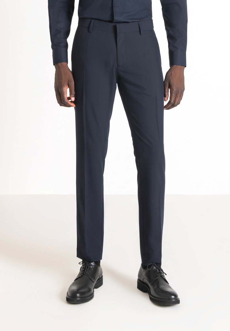 Pantaloni da abito navy, vestibilità tailored, tessuto morbido, frontale piatto, tasche laterali, passanti per cintura, abbinati a scarpe in pelle nera.