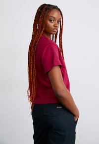 Bordeauxfarbene Baumwoll-T-Shirt mit kurzen Ärmeln und Rundhalsausschnitt. Getragen mit schwarzen Hosen, zeigt ein Seitenprofil des Models.