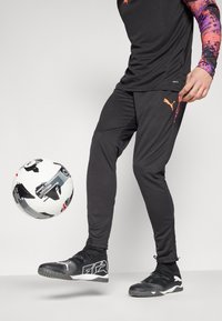 Black athletic pants with tapered fit, orange accents, paired with black sports shoes. A white patterned soccer ball is in motion.