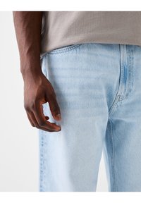 Närbild av en mörkhyad hand som vilar bredvid ljusblå jeans och en grå skjorta.