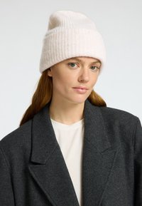 Beanie bianco a coste con design slouchy indossato sopra un cappotto grigio e una camicia chiara. Texture liscia, aspetto morbido.