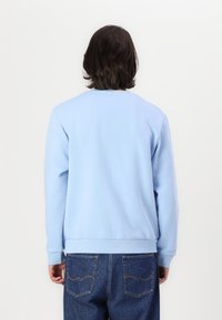 Person med axellångt mörkt hår som bär en ljusblå sweatshirt och mörkblå jeans, vänd bort mot en plain vit bakgrund.