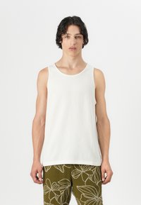 Top alb cu un model nervurat, cu decolteu rotund, purtat peste pantaloni verzi cu model. Materialul pare moale și ușor texturat.