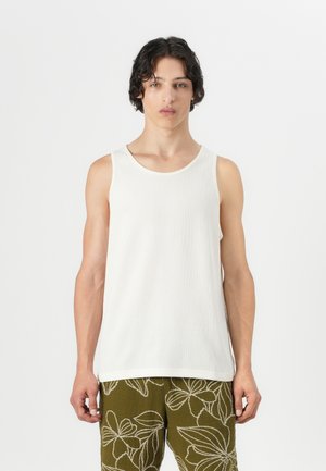 Weißes geripptes Tanktop mit rundem Ausschnitt, getragen über grünen gemusterten Hosen. Der Stoff wirkt weich und leicht strukturiert.