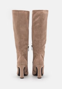 Par de botas de tacón altas de ante taupe con cremalleras laterales, mostradas desde atrás sobre un fondo blanco.