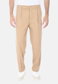Pantaloni beige a pieghe con una texture liscia, chiusura con zip e bottone, abbinati a sneakers bianche, presentano una vestibilità comoda.