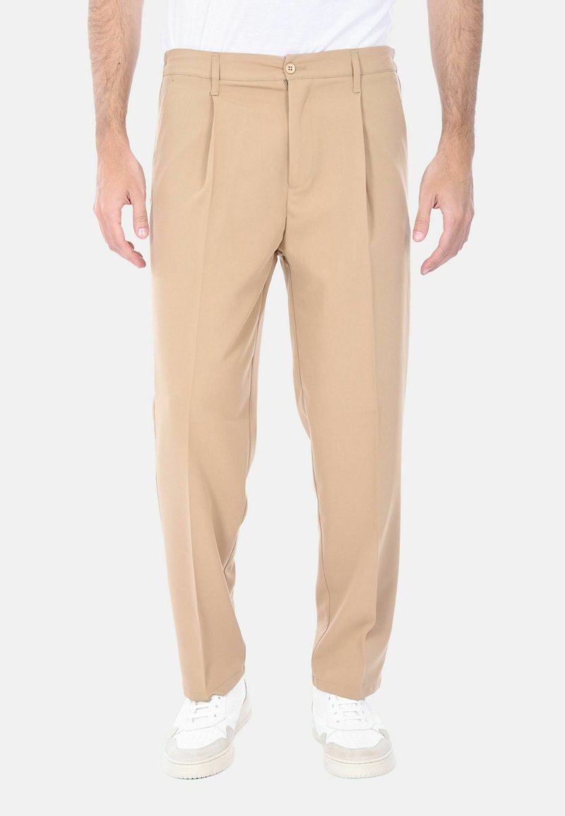 Pantaloni beige a pieghe con una texture liscia, chiusura con zip e bottone, abbinati a sneakers bianche, presentano una vestibilità comoda.