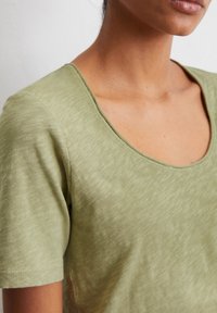 T-shirt verde oliva a maniche corte con scollatura ampia. Il tessuto ha un aspetto morbido e testurizzato con leggere variazioni di colore.