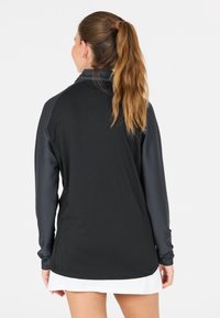 Kvinna med långt hästsvans som bär en svart och grå långärmad topp och en vit kjol, vänd bort från kameran mot en neutral bakgrund.