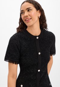 Cardigan nero in maglia strutturata con maniche corte, dettagli con frange e chiusure con bottoni argentati. Presenta una scollatura tonda e un design aderente.